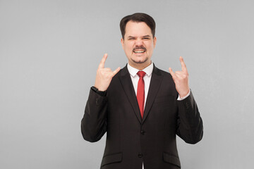 I'ts rock and roll, yeah! Expressive cool businessman showing rock sign. indoor studio shot. isolated on gray background. businessman with black suit, red tie and mustache looking at camera.