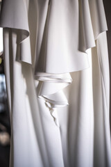 The perfect white silk wedding dress hanging in the room of the bride. Elegant and modern. Background. Close up of texture with shadows and layers.