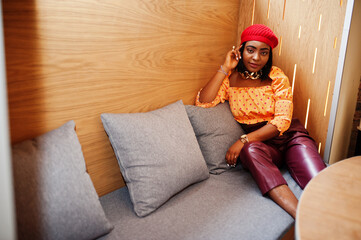 Elegant afro american woman in red french beret, big gold neck chain polka dot blouse and leather pants pose indoor.