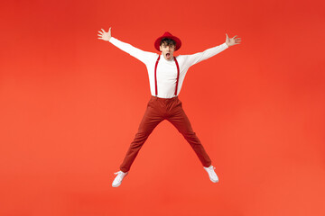 Fototapeta na wymiar Full length of young spanish latinos overjoyed weird shocked fun happy man 20s in hat white shirt trousers, suspenders jump high with outstretched hands legs isolated on red background studio portrait