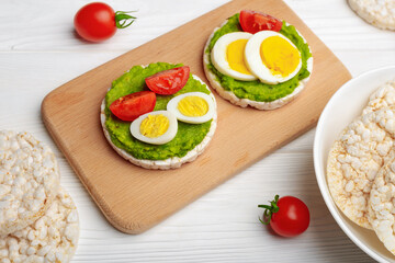 Fototapeta na wymiar Rice cakes with avocado mash and eggs with tomato on cutting board and white wooden background