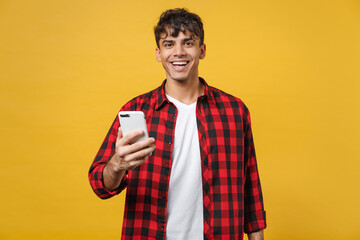 Young spanish latinos smiling handsome fashionable man 20s wearing red checkered shirt holding mobile cell phone chatting with friends in social network isolated on yellow background studio portrait.