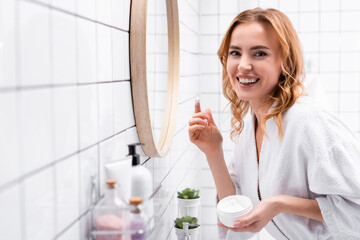 smiling woman holding jar with face cream near mirror near bottles on blurred foreground