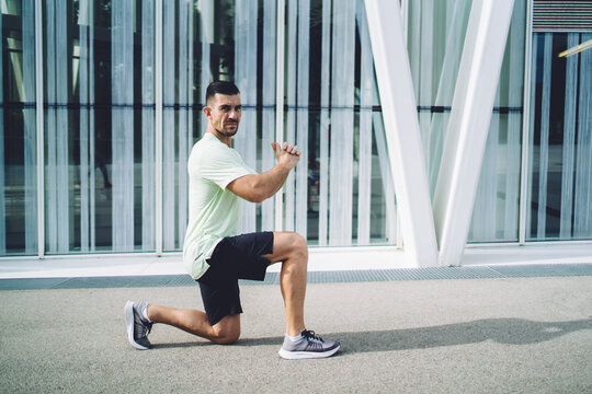 Portrait of determined male athlete stretching legs during morning training in urban city, muscular Caucasian jogger in sportswear concentrated on intensity practice doing squats at street