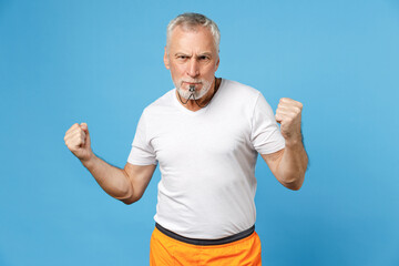 Elderly gray-haired sportsman trainer instructor angry strict man 50s in sportswear white t-shirt blow whistle clench fist isolated on blue background studio portrait. Fitness sport lifestyle concept.