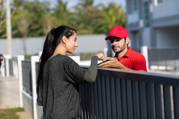 Signing signature on smart phone device to get a package. Beautifu woman receiving package from delivery man in red uniform at home.