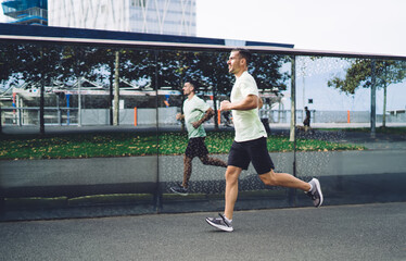Fototapeta premium Side view of determined male athlete getting ready to marathon have cardio morning training at city urbanity, motivated man in sportswear jogging with speed around street keeping healthy lifestyle
