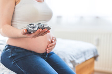 Happy pregnancy concept. Mother in blue jeans is holding baby shoes, tummy in the blurry background