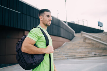 Pensive male bodybuilder with sportive backpack for gym stuff thoughtful looking away during morning time for cardio training, pondering Caycasian jogger standing at urban setting and thinking