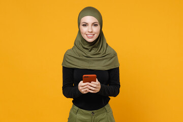 Smiling young arabian muslim woman wearing hijab black green clothes using mobile cell phone typing sms message isolated on yellow color background studio portrait. People religious lifestyle concept.