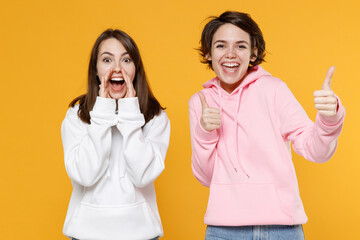 Excited two young women friends 20s in casual white pink hoodies standing scream news with hands near mouth showing thumbs up like gesture isolated on bright yellow color background studio portrait.