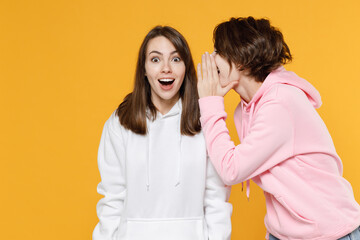 Shocked excited two young women friends 20s in casual white pink hoodies whispering gossip and tells secret behind her hand, sharing news isolated on bright yellow color background studio portrait.