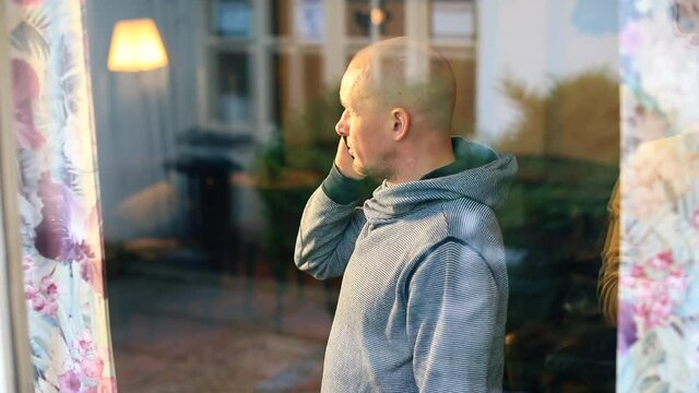 Bald man talking on the phone in a cafe. Window he puts the device to his ear, speaks a voice message. Shooting through glass, glare, reflections.