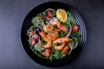 Salad with seafood, arugula, tomatoes, cucumbers, red onion and lemon on a black plate. Black background, close-up.