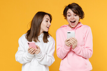 Excited amazed smiling two young women friends 20s wearing casual white pink hoodies standing using mobile cell phone typing sms message isolated on bright yellow color background studio portrait.