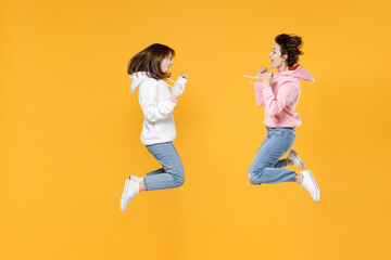 Fototapeta na wymiar Full length side view of happy two young women friends 20s wearing basic white pink hoodies jumping doing winner gesture celebrating say yes isolated on bright yellow color background studio portrait.
