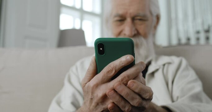 Close up view of senior male retiree chatting with family while typing at phone sreen.Positive bearded man with grey hair reading message and smiling while sitting on sofa at home