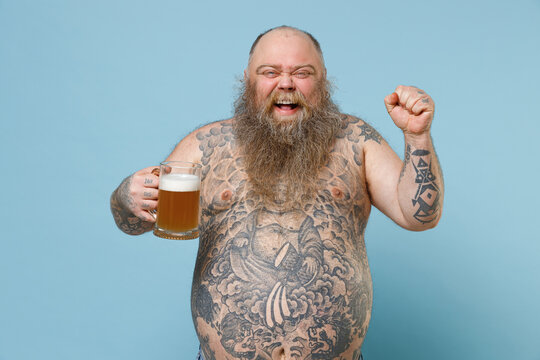 Joyful fat pudge obese chubby overweight man has tattooed naked bare big belly hold glass of beer doing winner gesture isolated on blue background studio. Weight loss obesity unhealthy diet concept.