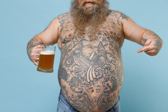 Cropped image fat pudge obese chubby overweight man has tattooed big belly hold glass of beer pointing index finger on stomach isolated on blue background. Weight loss obesity unhealthy diet concept.