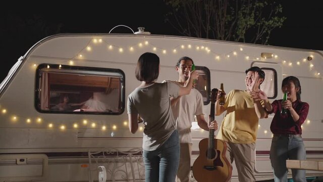 Friends enjoying outdoor guitar party at night, clink and drink beer.