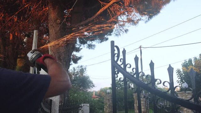 Caucasian Man is Busy Using Electric Grinder Tool to Smooth an Iron Gate. Fixing the Entrance Door of a House with Pine Trees. 4K Video Footage