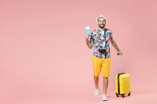 Full length of cheerful young traveler tourist man in summer clothes hat hold suitcase passport tickets isolated on pink background studio. Passenger traveling on weekends. Air flight journey concept.