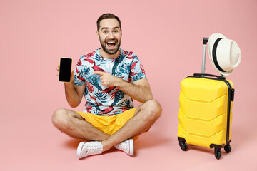 Full length excited traveler tourist man sit on floor point index finger on mobile phone with blank empty screen isolated on pink background. Passenger travel on weekend. Air flight journey concept.
