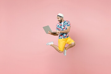 Full length of excited young traveler tourist man in summer clothes hat jumping working on laptop computer isolated on pink background. Passenger traveling on weekend. Air flight journey concept.