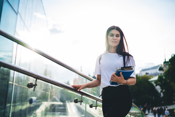 Half length portrait of attractive female schooler with education equipment in hand posing at urban...