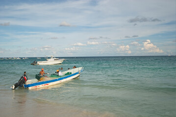 On the seashore, fishermen on their boat are cleaning fish; in the background is a cloudy sky and tourists are playing in the water. Mexican Caribbean
