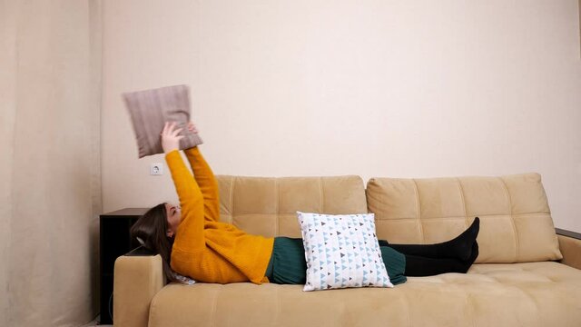 Bored unemployed young woman in yellow sweater lies on brown sofa and throws pillows from idleness catching near white wall at home