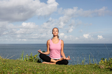 Fototapeta na wymiar young beautiful smiling blonde woman in sportswear doing yoga sitting on the grass with blue sea and sky on the background