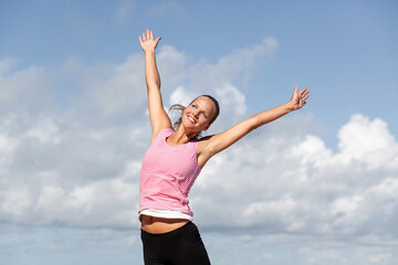 young beautiful happy smiling woman in sportswear spread her arms to the sides against the background of the cloudy sky