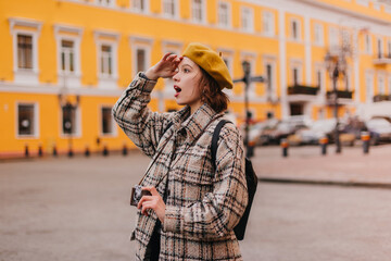 Portrait of woman in yellow beret with retro camera in her hands. Girl in plaid coat looks at magnificent European buildings with surprise and delight