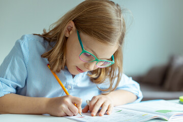 Pretty teen schoolgirl in glasses making homework at desk at home during pandemic distance learning