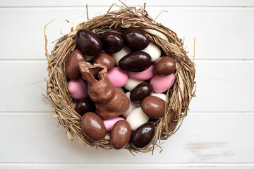 Top view of bird nest with chocolate bunny and sweet easter eggs on white wooden table
