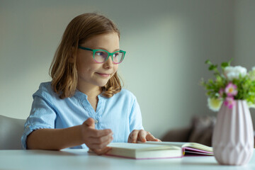 Fototapeta na wymiar Pretty teen girl reading book at desk during pandemic distance learning