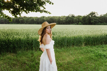 Beautiful young woman in a boho style dress and hat on a field with wheat at sunset.