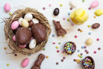 Top view of bird nest with chocolate easter eggs, chocolate bunny and sweets on white wooden table