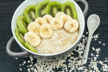 Oatmeal porridge with bananas, kiwi in a bowl on a dark background close-up. Healthy breakfast.