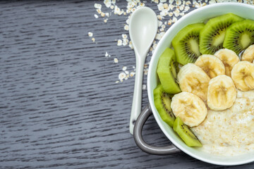 Oatmeal porridge with bananas, kiwi in a bowl with a spoon on a dark background with copy space, top view. Healthy breakfast.