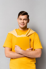 Cheerful student in yellow clothes holding notebook on grey background