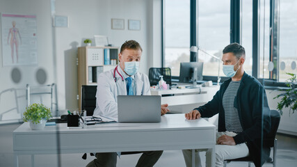 Family Doctor in Protective Mask is Reading Medical History of Young Male Patient and Speaking with Him During Consultation in a Health Clinic. Physician in Lab in Front of Computer in Hospital Office