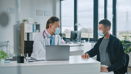Family Doctor in Protective Mask is Reading Medical History of Young Male Patient and Speaking with Him During Consultation in a Health Clinic. Physician in Lab in Front of Computer in Hospital Office
