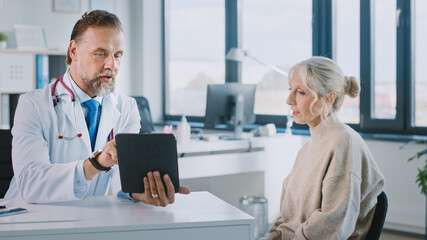 Obraz premium Friendly and Cheerful Family Doctor is Reading Medical History of Senior Female Patient During Consultation in a Health Clinic. Physician Using Tablet Computer in Hospital Office.