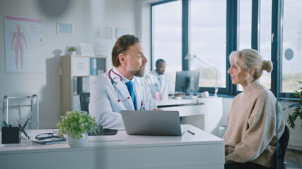 Friendly and Cheerful Family Doctor is Reading Medical History of Senior Female Patient During Consultation in a Health Clinic. Physician Using Laptop Computer in Hospital Office.