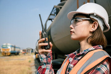 Portrait of beautiful woman engineering using walkie talkie and with wear hardhat in front of train station. Behind the scenes, workers and employees are filling up and repairing the train.