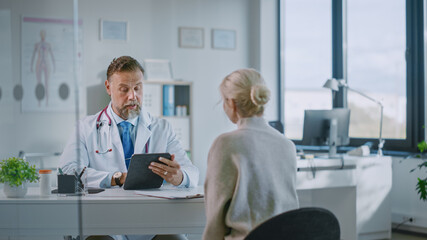 Fototapeta premium Friendly and Cheerful Family Doctor is Reading Medical History of Senior Female Patient During Consultation in a Health Clinic. Physician Using Tablet Computer in Hospital Office.