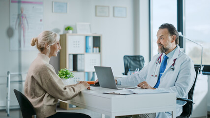 Fototapeta premium Friendly and Cheerful Family Doctor is Reading Medical History of Senior Female Patient During Consultation in a Health Clinic. Physician Using Laptop Computer in Hospital Office.
