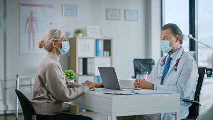 Obraz premium Family Doctor in Protective Mask is Reading Medical History of Senior Female Patient and Speaking with Her During Consultation in a Health Clinic. Physician in Lab in Front of Computer in Hospital.